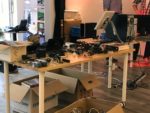 Ein Tisch im Vordergrund, andere im Hintergrund. Darauf viele technische Geräte, Monitore, Kabel. Darunter Kartons mit weiterem Material.