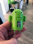 Ein grüner runder Robotor mit zwei Antennen und roten Augen, als Füße die beiden Stecker für die Steckdose.