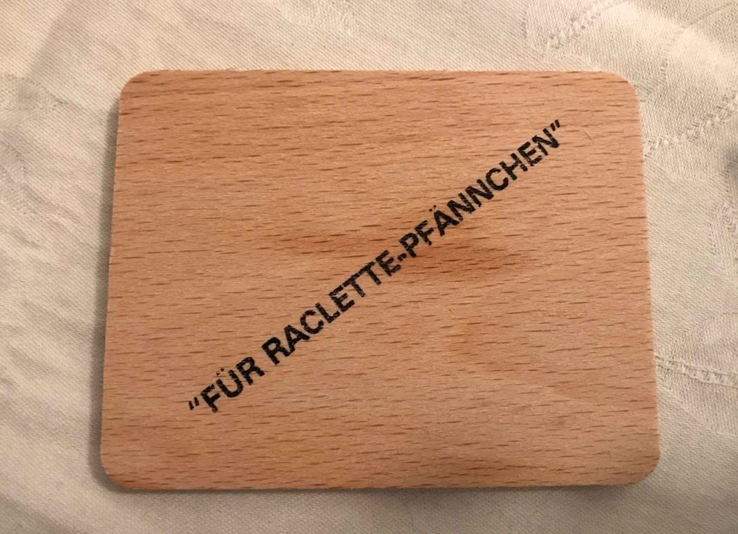 Ein kleines Holzbrett mit Fettflecken, darauf ist schräg in Anführungszeichen gedruckt: Für Raclette-Pfännchen