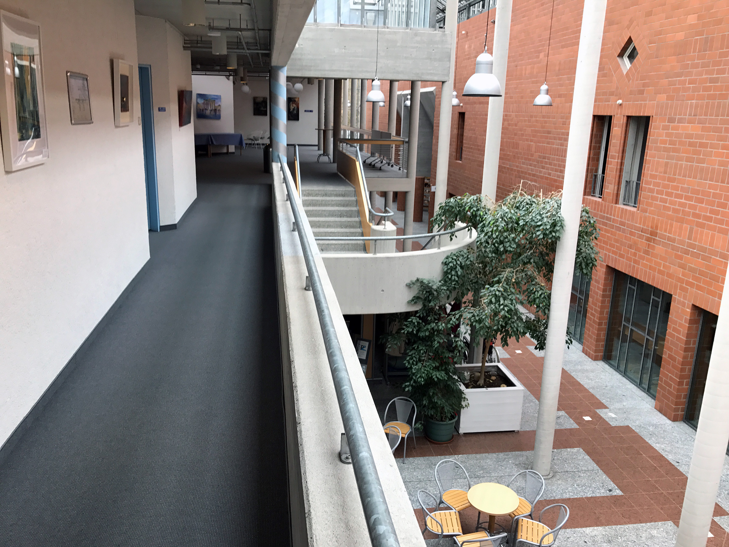 Der Blick auf von der Galerie des ersten Stocks, links eine Wand, rechts blickt man nach unten. Viel Beton und eine Ziegelwand.