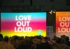 Aus den hinteren Reihen eines großen dunklen Saals die Bühne mit einer bunten Leinwand, auf der "Love out loud" steht, daneben ein gleich gestaltetes großes Plakat.