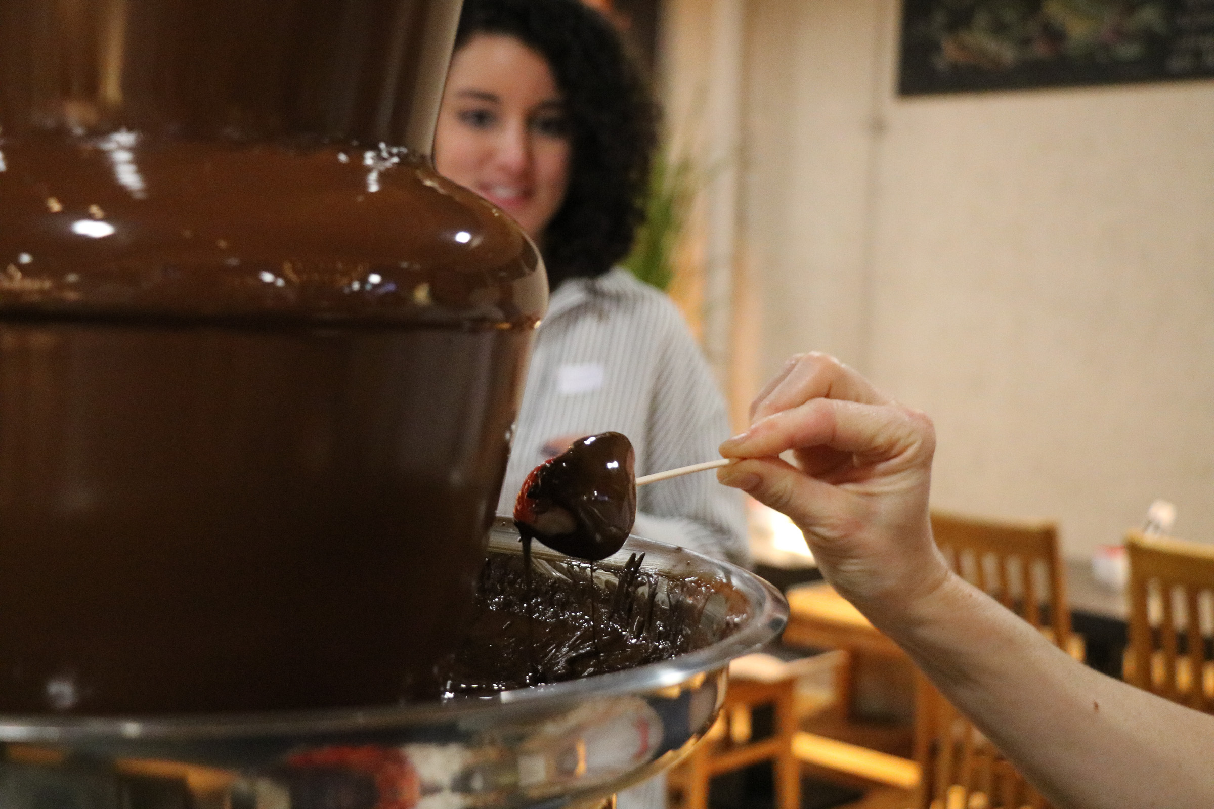 Eine Person hält eine Erdbeere an einem Holzstab unter flüssige Schokolade. Im Hintergrund eine Frau, die interessiert zusieht.
