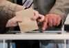 Zwei Hände halten einen Brief über den Einwurfschlitz einer Wahlurne. Dahinter zeigt eine Hand auf den Schlitz.
