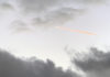 Ein Flugzeugkondensstreifen, der durch die Abendsonne rot-orange vor dem grauen Himmel leuchtet.