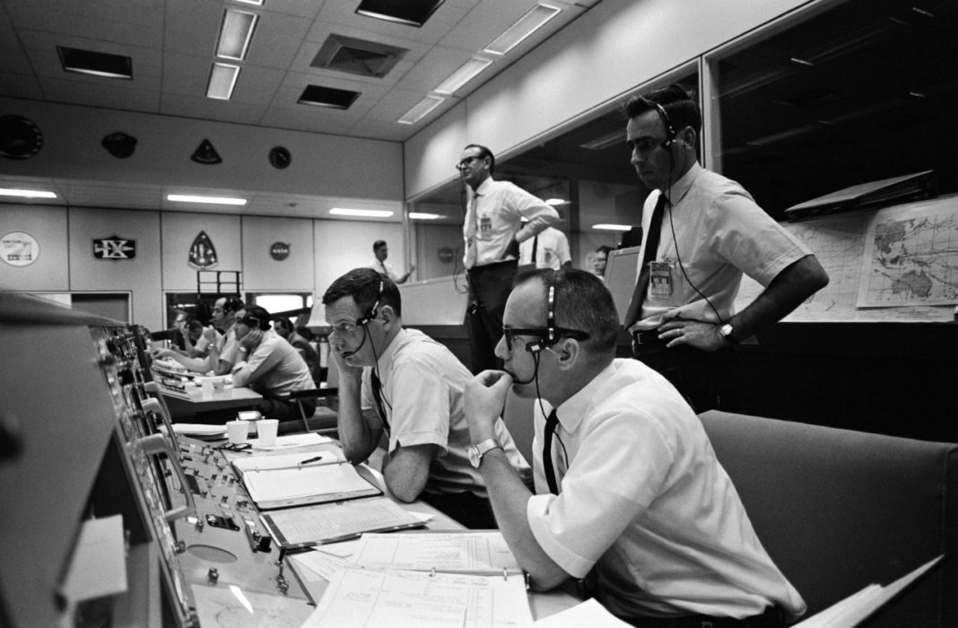Männer im Look der 1960-er mit Headsets sitzen an einem Schaltpult bzw. stehen dahinter. Sie alle blicken in die gleiche Richtung aus dem Bild hinaus.