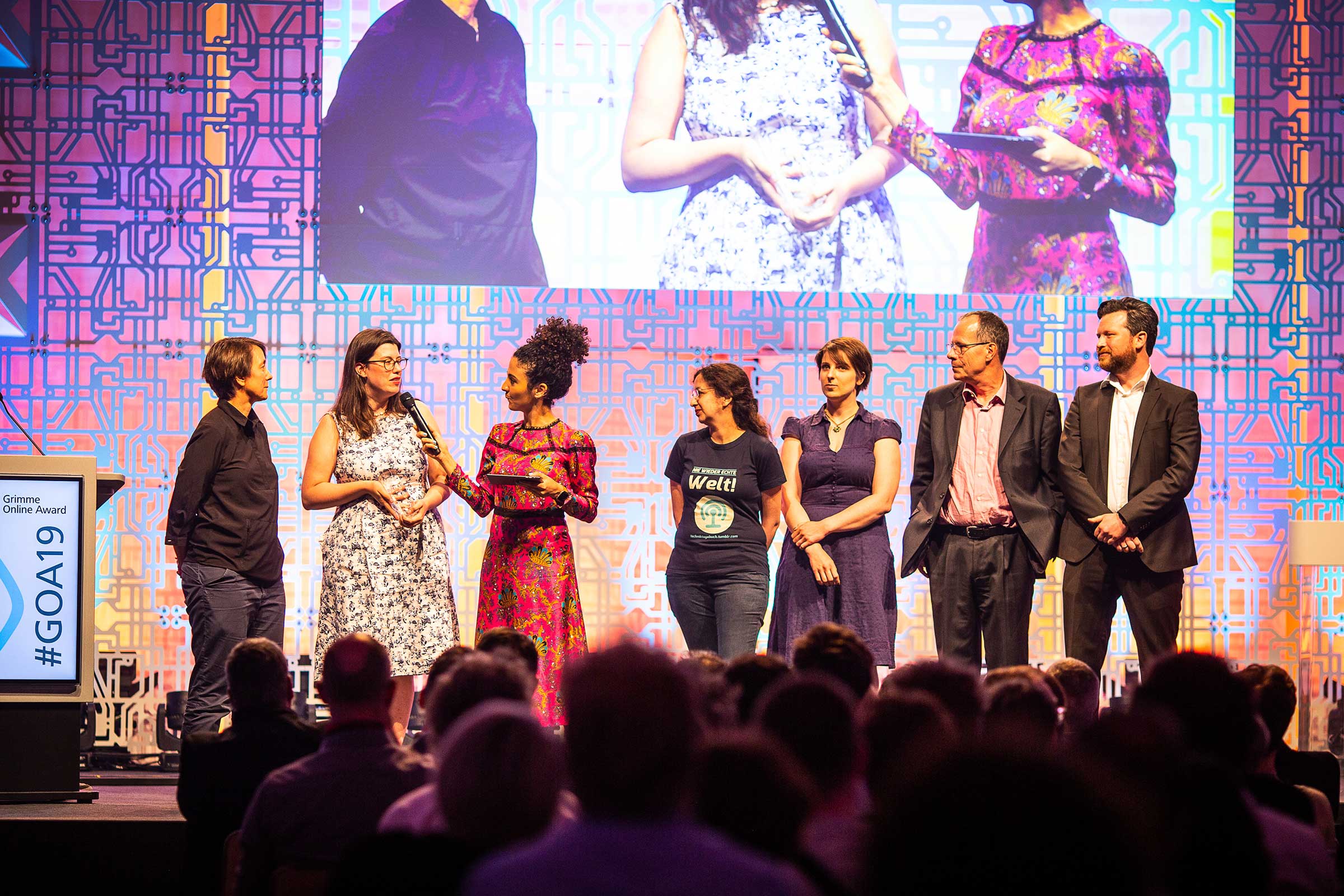 Sieben Menschen auf einer Bühne, von links nach rechts: fünf Frauen, zwei Männer, ganz rechts ich. Die Moderatorin (dritte von links) hält der zweiten von links ein Mikrofon vor das Gesicht.
