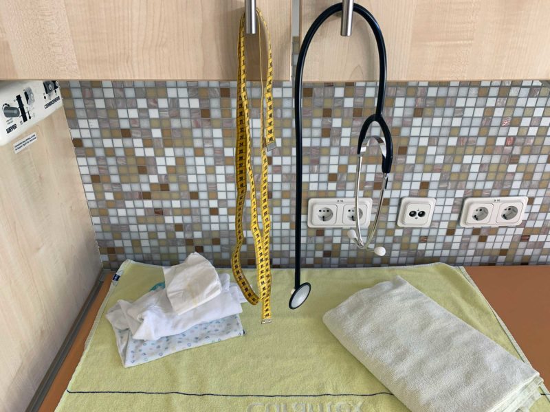 Bild aus dem Kreißsaal: Vom Hängeschrank hängen ein Maßband und ein Stethoskop, unterhalb eine Wickelauflage mit bereitgelegten Klamotten für das Baby und einem Handtuch.