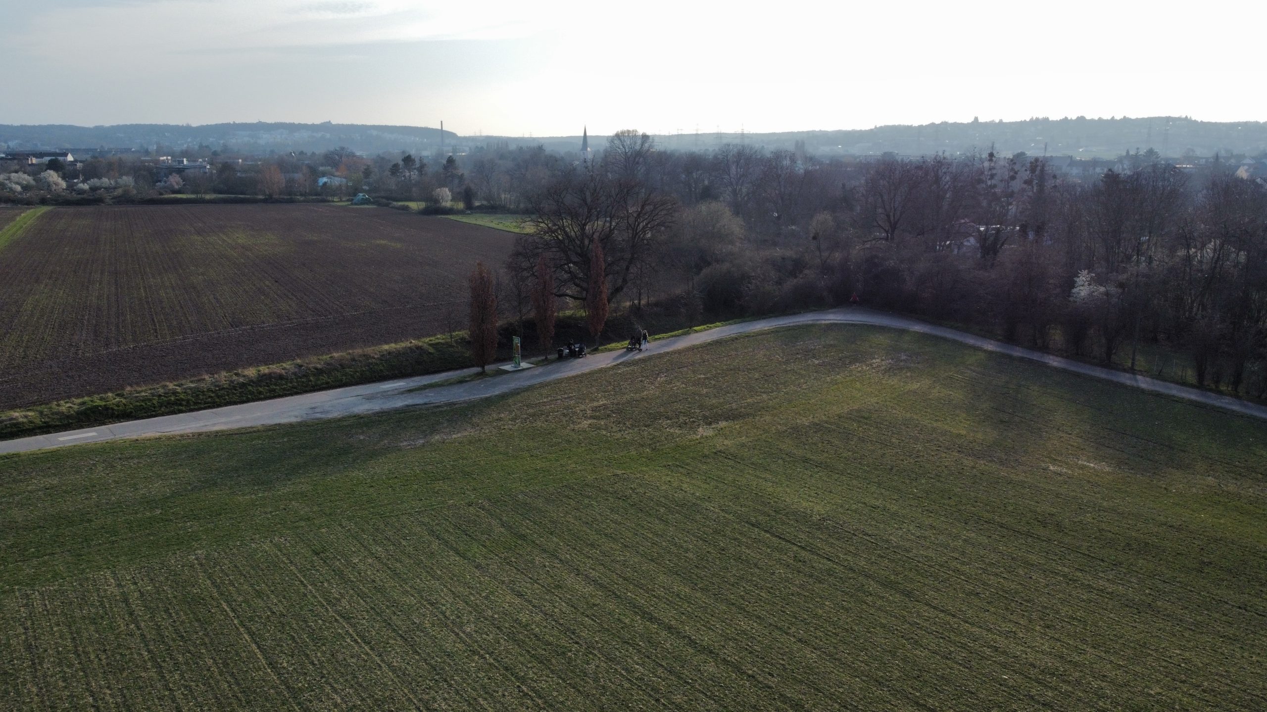 Blick von oben auf Felder, dazwischen ein Weg, am Horizont Häuser und eine hügelige Landschaft.