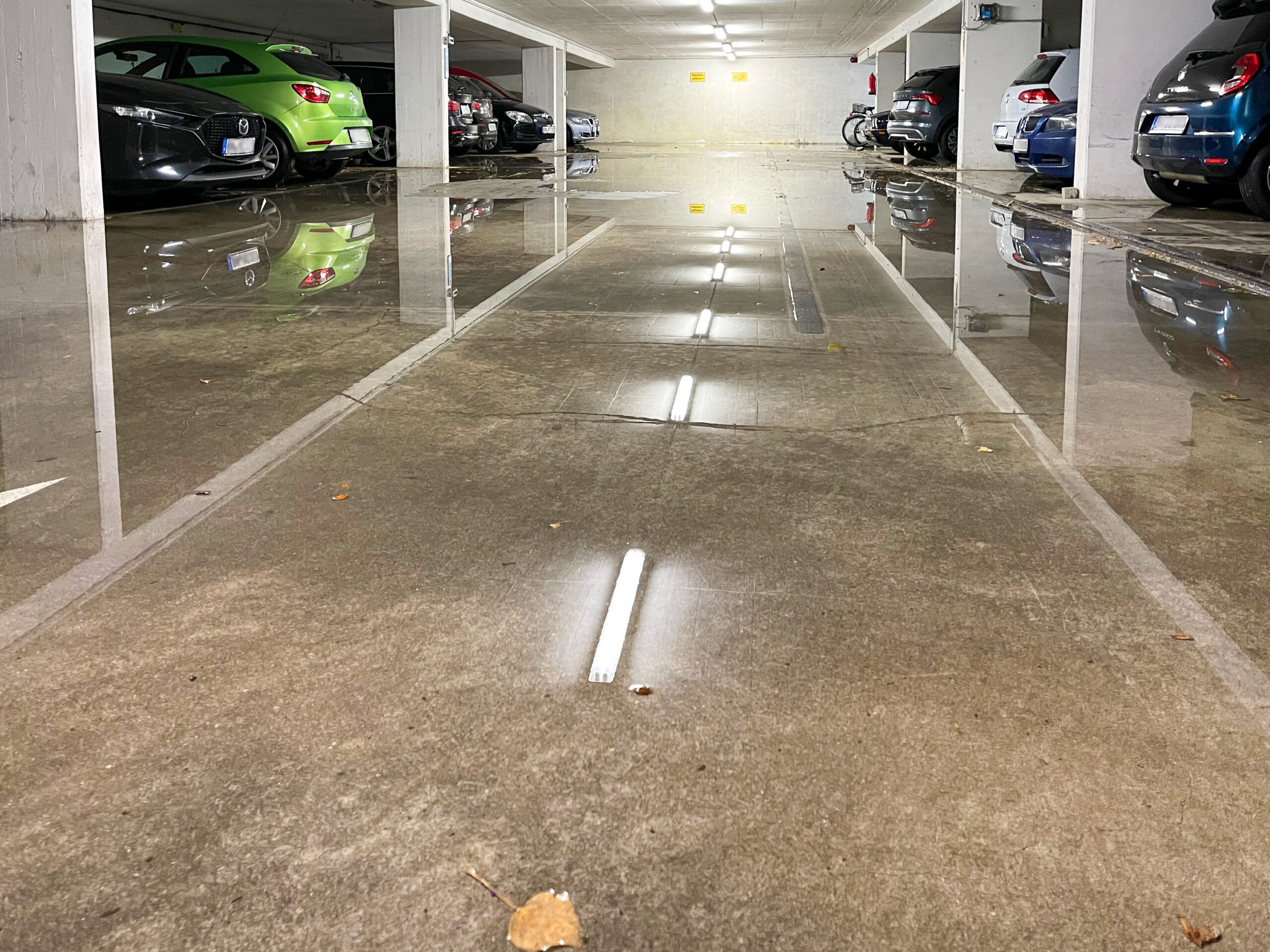 Eine Tiefgarage, links und rechts parken Autos, in der Mitte steht etwa zwei Zentimeter hoch das Wasser, sodass sich Autos und Beleuchtung spiegeln.