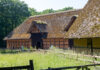 Ein altes Fachwerkhaus mit Reetdach und gelben Wänden, davor eine lang gewachsene Wiese mit Holzumzäunung.