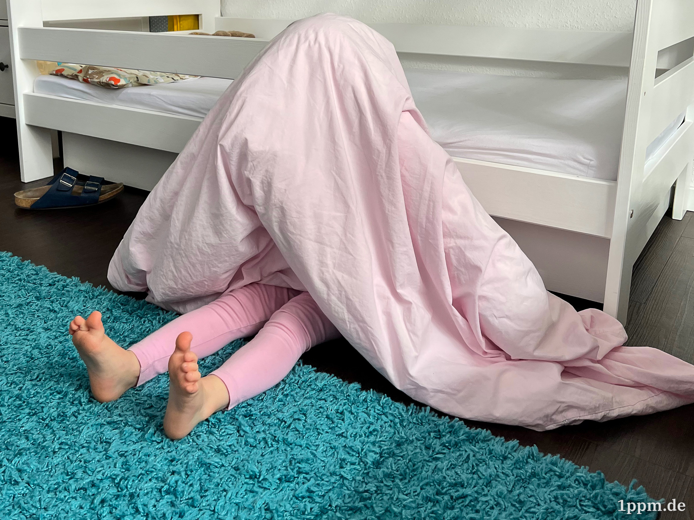 Klein-Lea sitzt auf dem Boden an ihr Bett gelehnt und hat sich ihre Decke über den Kopf gezogen. Man sieht nur ihre Beine herausragen.
