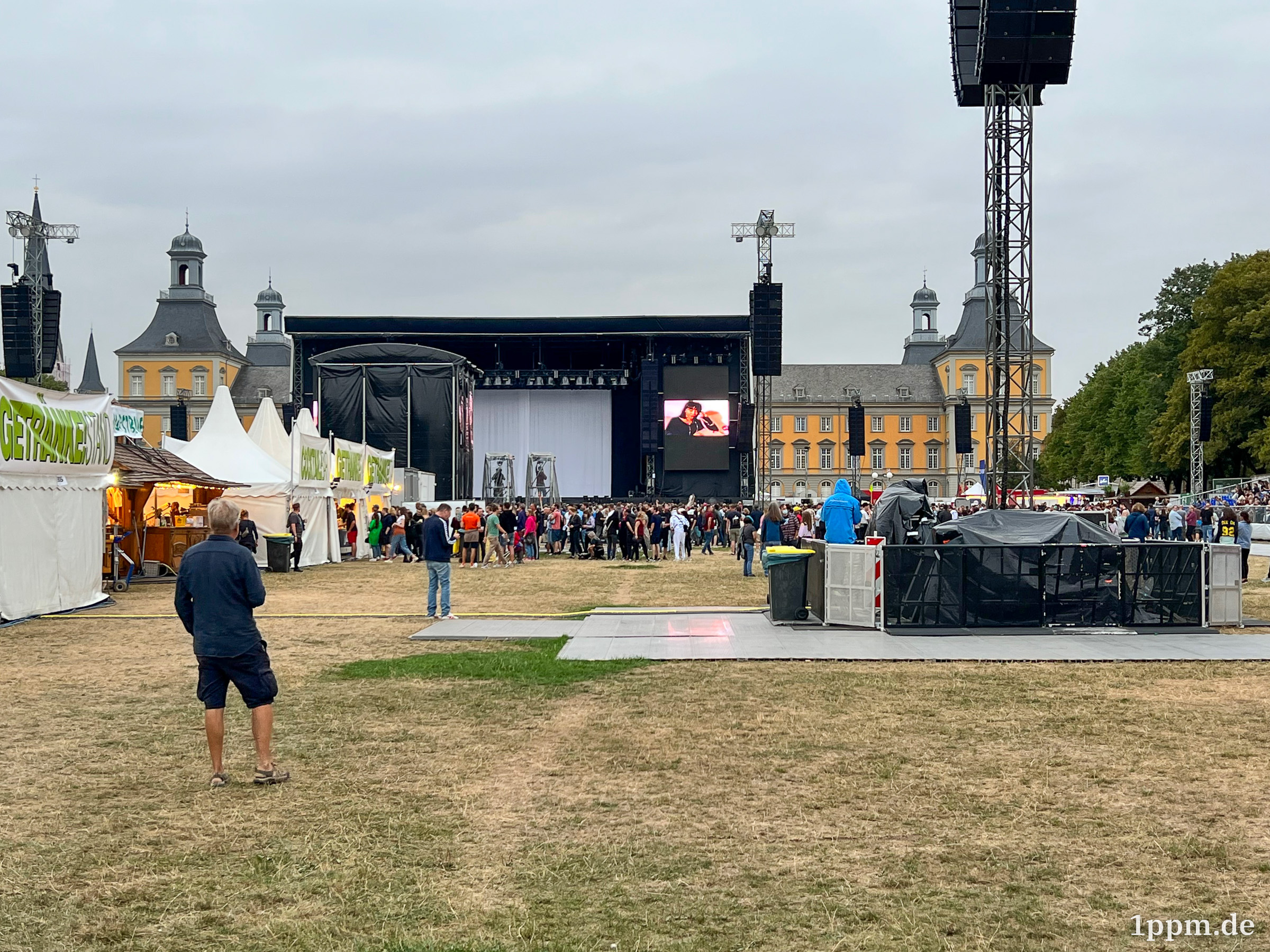 Eine Wiese mit Bühne und Getränkeständen, hinter der Bühne sieht man das gelbe Uni-Bonn-Gebäude