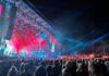 Eine Bühne von schräg links mit rot-blau leuchtenden Scheinwerfern, davor viele, viele Menschen