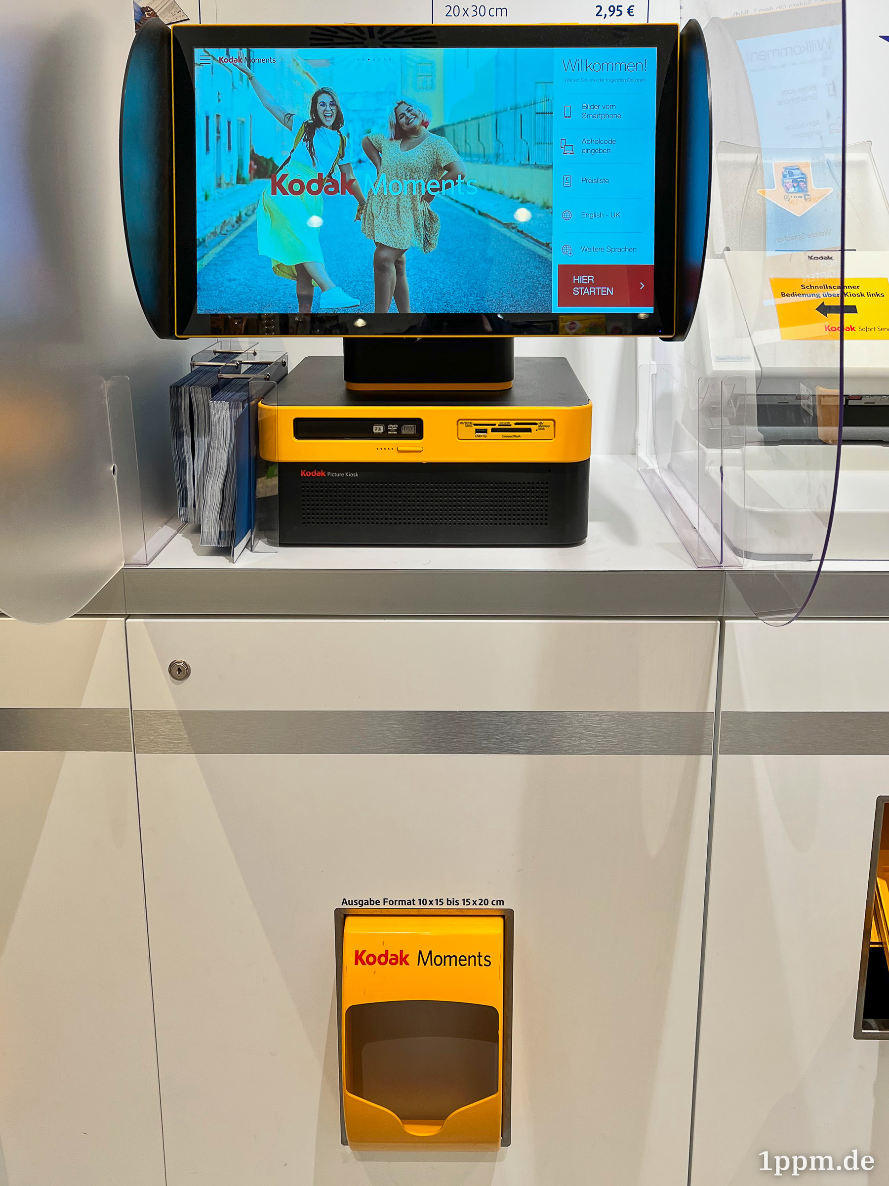 Ein großer Bildschirm auf einem gelb-schwarzen Kasten steht auf einem Schrank. Der Schrank hat unten ein gelbes, mit „Kodak Moments“ beschriftetes Ausgabefach.