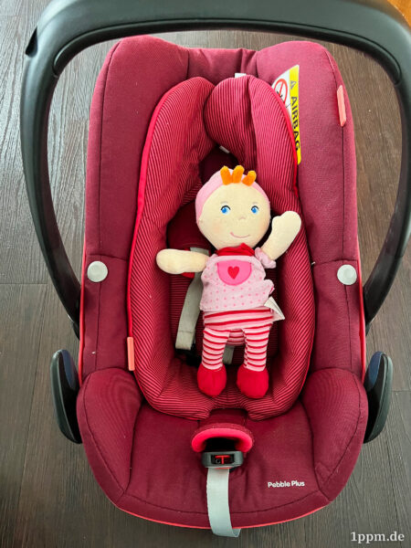 Eine Puppe liegt im Maxi-Cosi und winkt.