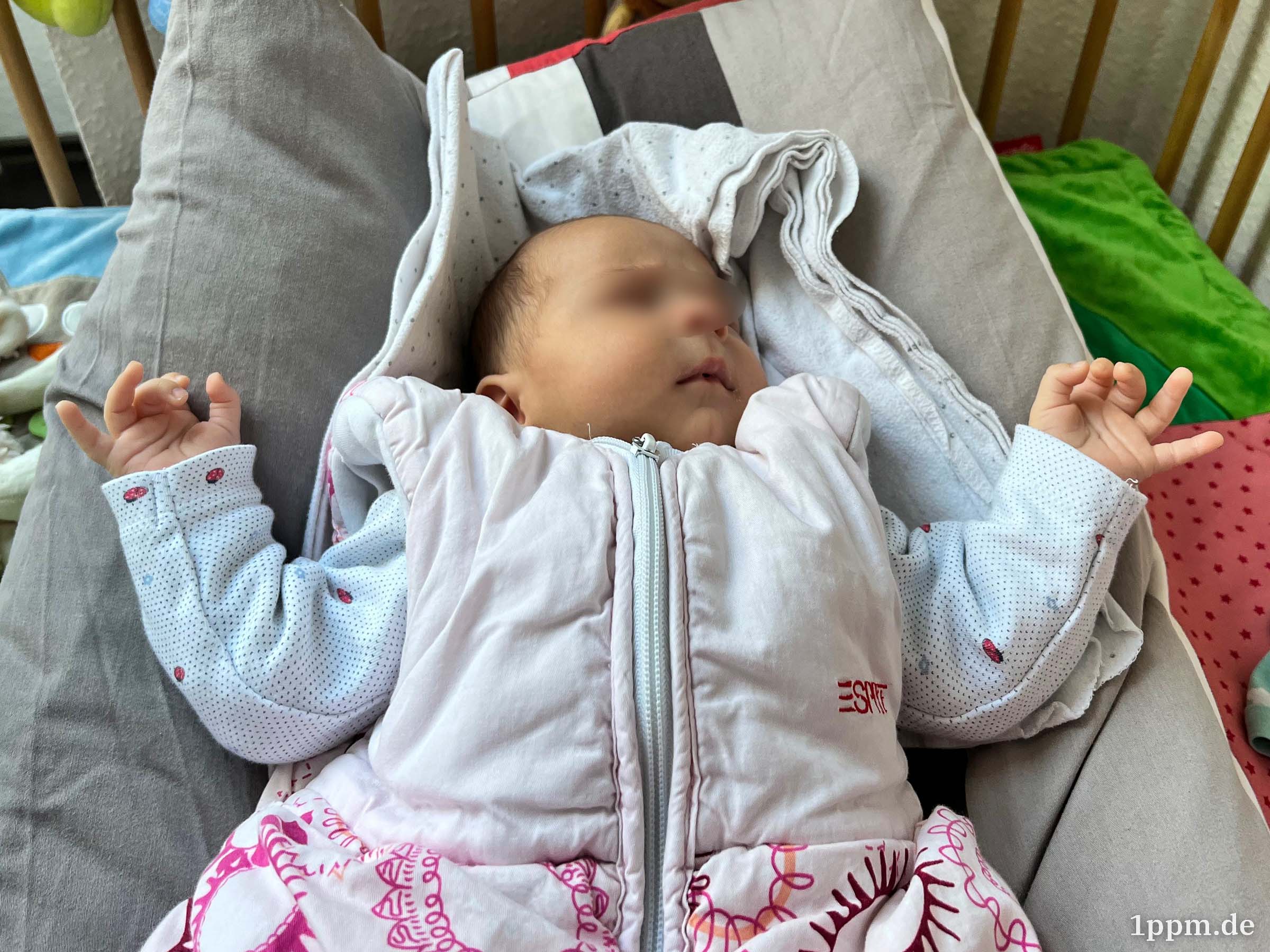 Ein Baby im Schlafsack liegt auf einem Kissen und hält beide Hände leicht nach oben.