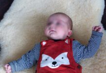 Ein Baby liegt auf einem Fell auf dem Boden. Es trägt einen roten Strampelanzug, auf dem ein Fuchskopf abgebildet ist.