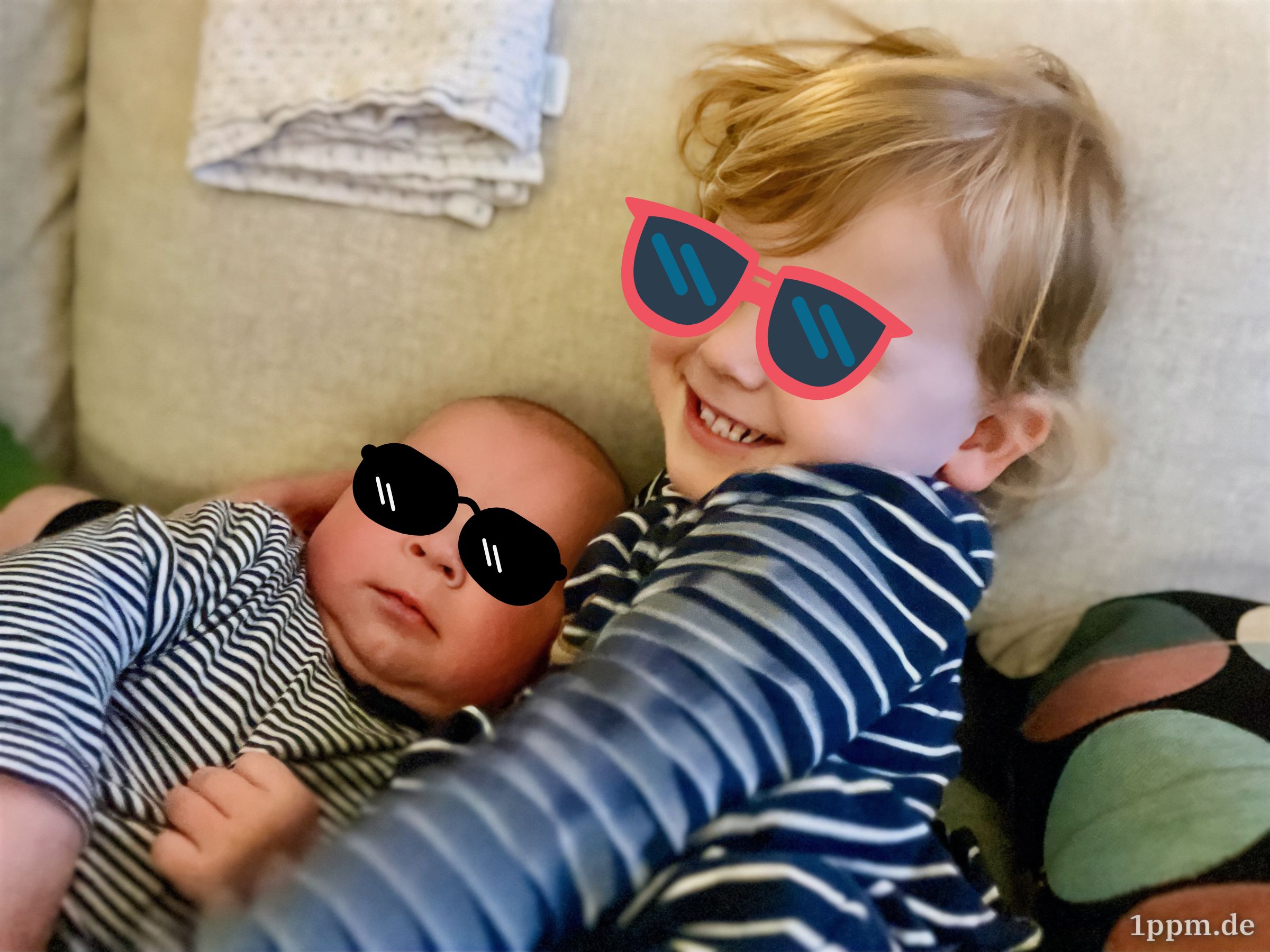 Der Babysohn und die Kleinkindtochter nebeneinander auf dem Sofa, er schmiegt sich scheinbar an sie, sie lacht ganz stolz. Auf beiden Augen wurden Sonnenbrillen retuschiert.
