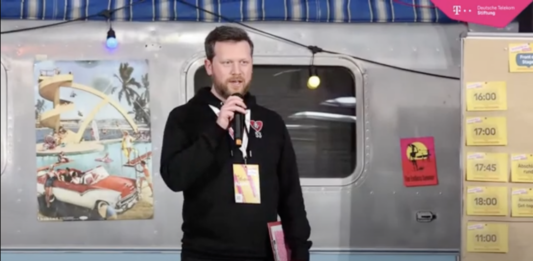 Johannes im schwarzen Hoodie, in der einen Hand ein Mikrofon, in der anderen ein Klemmbrett.