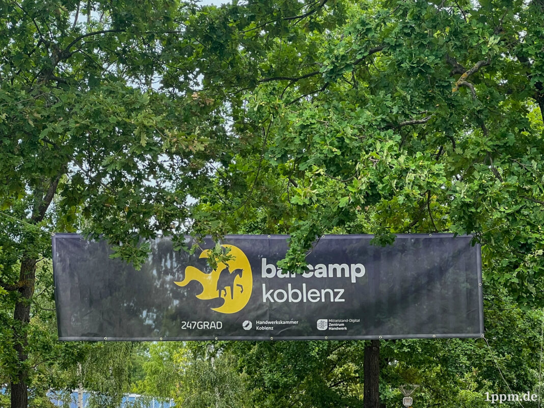 Ein Banner mit dem Logo vom Barcamp Koblenz hängt zwischen Bäumen.