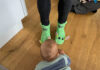 Ein Baby von hinten, das auf dem Boden krabbelt. Vor ihm zwei Beine in schwarz, die in zwei giftgrünen Socken enden, die jeweils zwei Augen und Ohren haben. Sie sollen Grogu aus Star Wars darstellen.