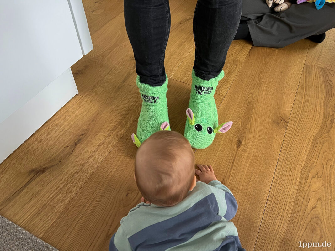 Ein Baby von hinten, das auf dem Boden krabbelt. Vor ihm zwei Beine in schwarz, die in zwei giftgrünen Socken enden, die jeweils zwei Augen und Ohren haben. Sie sollen Grogu aus Star Wars darstellen.