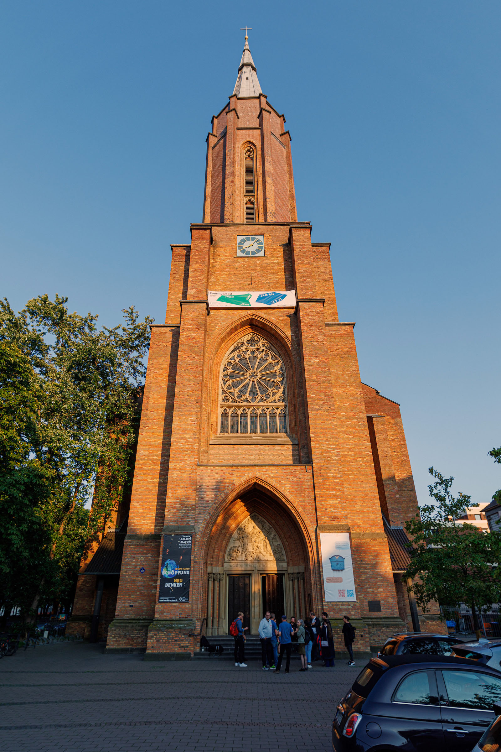 Ein hoher Kirchturm aus roten Sandsteinen, der in der Abendsonne leuchtet. Klein davor eine Gruppe Menschen.