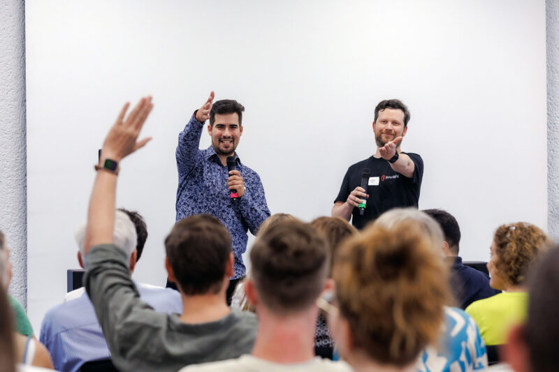 Sascha und Marc zeigen von der Bühne auf eine Person im Publikum, die die Hand hebt. Das Publikum sieht man nur von hinten.