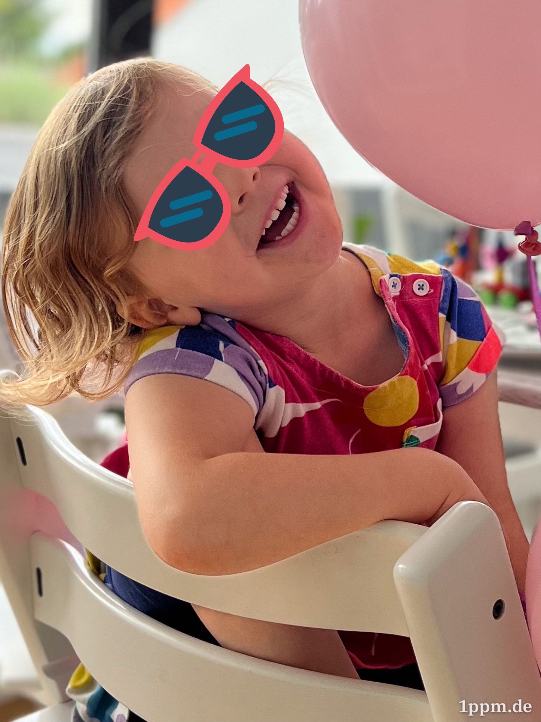Ein Mädchen (mit retuschierter Sonnenbrille) hängt mit einem Arm über der Stuhllehne und lacht. Im Hintergrund ein rosafarbener Luftballon.
