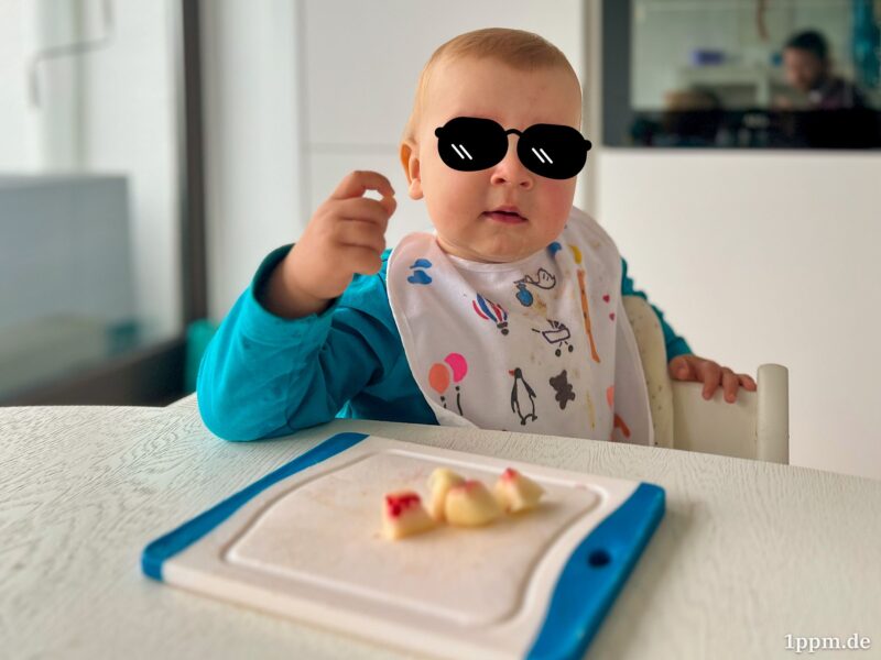 Klein-Tyler sitzt schräg am Tisch, den rechten Ellbogen aufgestützt. Mit der rechten Hand macht er mit Daumen und Zeigefinger ein O. Vor ihm liegt ein Bettchen mit Apfelstücken. Er guckt aufgeschlossen zum Fotografen, der sich im Hintergrund im Backofen spiegelt (es ist der Autor). Über Klein-Tylers Augen wurde eine Sonnenbrille retuschiert.