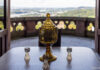 Eine Glaskaraffe aus gelblich-bräunlichen Glas, reich verschnörkelt, drumherum vier kleine Gläser in gleicher Farbe auf einem dunkelbraunen Tisch. Im Hintergrund ist ein Balkon zu erahnen, der den Blick auf das Rheintal und Bonn freigibt.