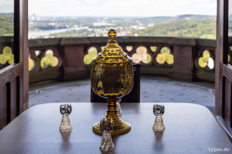 Eine Glaskaraffe aus gelblich-bräunlichen Glas, reich verschnörkelt, drumherum vier kleine Gläser in gleicher Farbe auf einem dunkelbraunen Tisch. Im Hintergrund ist ein Balkon zu erahnen, der den Blick auf das Rheintal und Bonn freigibt.