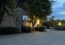Ein Pausenhof im Zwielicht mit zwei leuchtenden länglichen Laternen, daneben ein Basketballkorb
