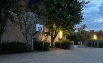 Ein Pausenhof im Zwielicht mit zwei leuchtenden länglichen Laternen, daneben ein Basketballkorb
