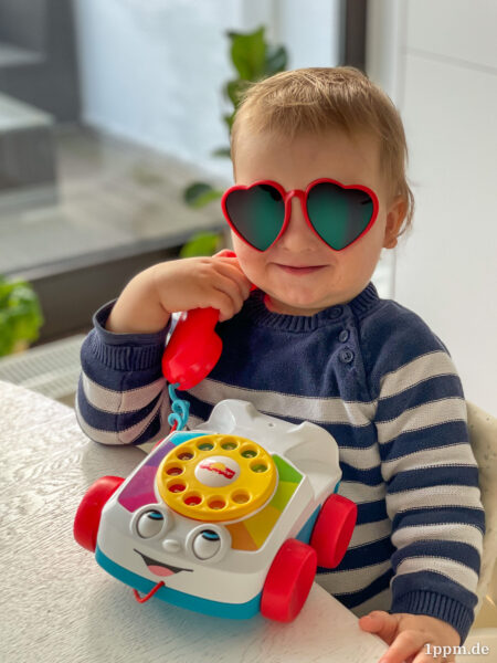Klein-Tyler am Spielzeugtelefon, er hält den Hörer in der Hand und blickt freudig in die Kamera. Auf seine Augen wurde eine auffällige Herzchen-Sonnenbrille retuschiert.