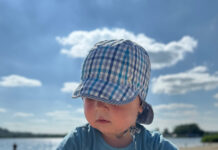 Kleines Kind in Hut und blauem Shirt sitzt am Strand.