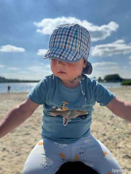 Kleines Kind in Hut und blauem Shirt sitzt am Strand.