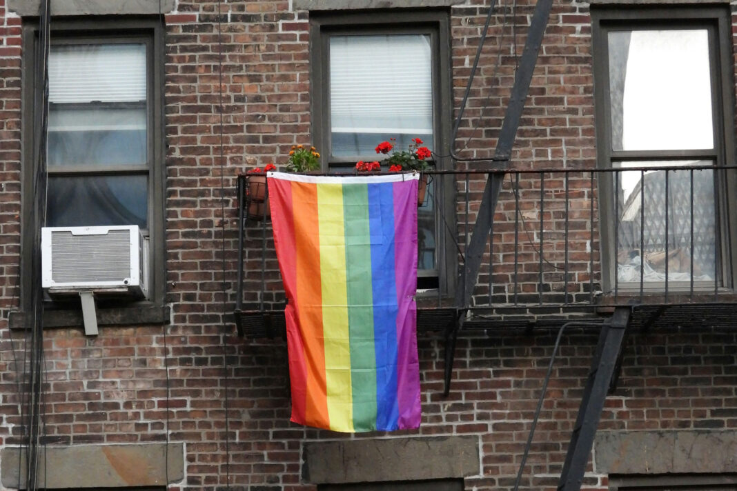 Eine Regenbogenflagge, die aus einem Fenster eines braun geziegelten Hauses hängt.