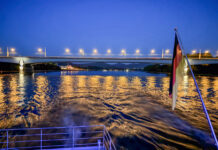 Hinten vom Schiff raus bei schon größerer Dunkelheit. Man erahnt eine Brücke, deren Lichter sich im Wasser spiegeln und sieht ansonsten viel Wasser und die Deutschlandfahne der Schiffsbeflaggung.