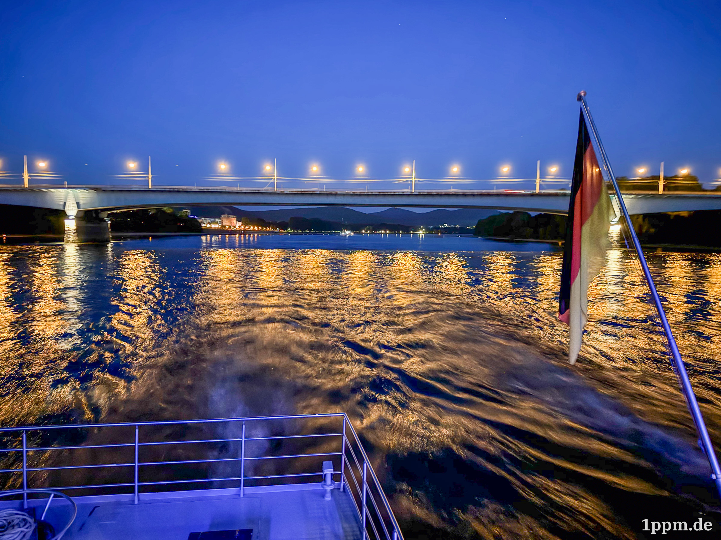 Hinten vom Schiff raus bei schon größerer Dunkelheit. Man erahnt eine Brücke, deren Lichter sich im Wasser spiegeln und sieht ansonsten viel Wasser und die Deutschlandfahne der Schiffsbeflaggung.