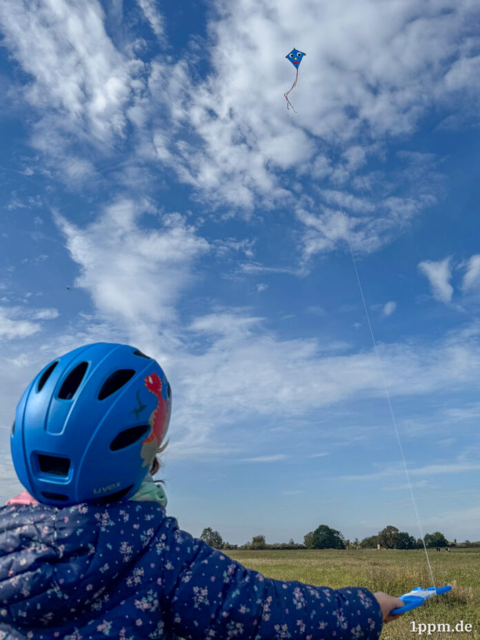 Ein Kind mit blauem Fahrradhelm von hinten, es hält einen blauen Drachen mit Gesicht an einer langen Schnur und schaut zu ihm in den Himmel.