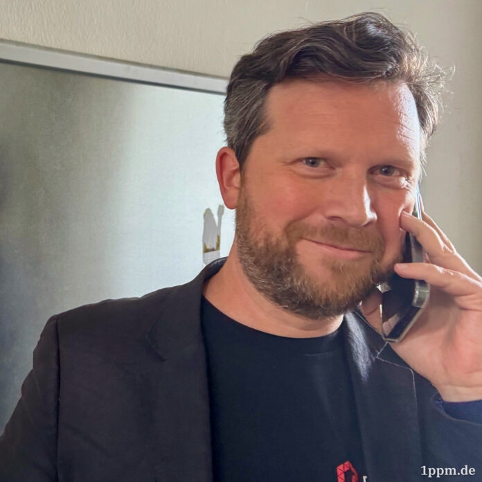 Ein Mann mit Bart hält sein Handy ans Ohr und lächelt in die Kamera.