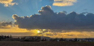 Ein Sonnenuntergang, teilweise verdeckt von einer dicken Wolke. Im Vordergrund erahnt man ein Feld, im Hintergrund Bebauung.