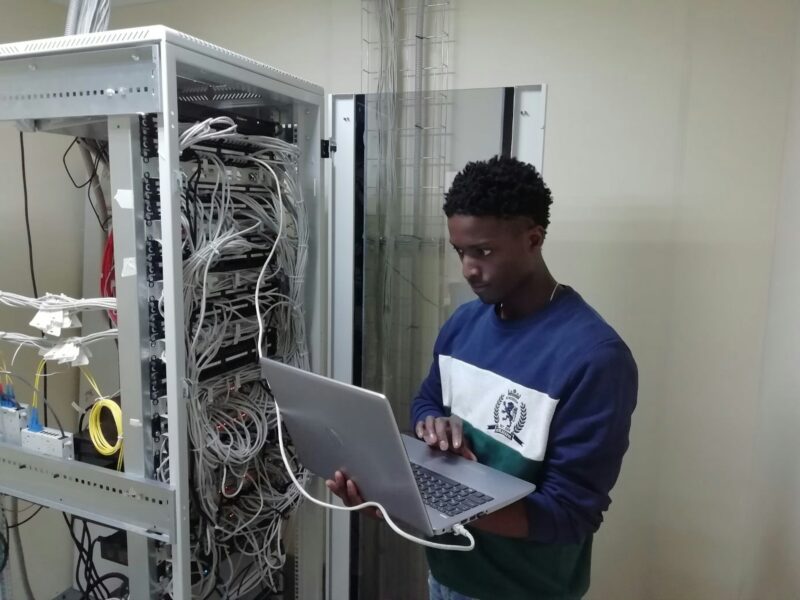 Mann in blauem Pullover der ein silbernes MacBook stehend in der Hand hält. Er steht neben einem Serverrack. Vom Computer führt ein Netzwerkkabel direkt an einen Server. Der Mann starrt konzentriert auf den Monitor.