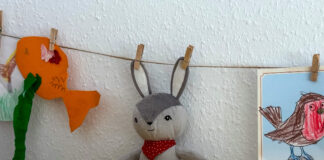 Ein kleiner Kuscheltierhase wurde mit Wäscheklammern an beiden Ohren an eine Schnur aufgehängt. Links daneben ein gebastelter, orangefarbener Fisch an der Schnur, rechts ein ausgemaltes Bild einer Meise (mit rotem Bauch).