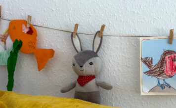 Ein kleiner Kuscheltierhase wurde mit Wäscheklammern an beiden Ohren an eine Schnur aufgehängt. Links daneben ein gebastelter, orangefarbener Fisch an der Schnur, rechts ein ausgemaltes Bild einer Meise (mit rotem Bauch).