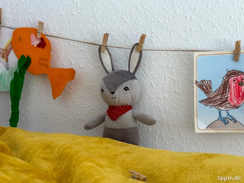 Ein kleiner Kuscheltierhase wurde mit Wäscheklammern an beiden Ohren an eine Schnur aufgehängt. Links daneben ein gebastelter, orangefarbener Fisch an der Schnur, rechts ein ausgemaltes Bild einer Meise (mit rotem Bauch).