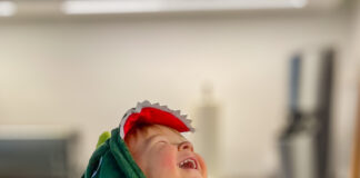 Ein Kind in einem Dinosaurier-Kostüm, das sich an der Küchenarbeitsplatte festhält und lauthals lacht und damit die Augen zusammenkneift und den Kopf in den Nacken wirft.