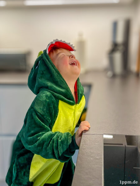 Ein Kind in einem Dinosaurier-Kostüm, das sich an der Küchenarbeitsplatte festhält und lauthals lacht und damit die Augen zusammenkneift und den Kopf in den Nacken wirft.