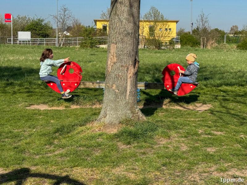 Zwei Kinder auf einer Wippe, ziemlich genau in der Mitte davor im Bild ein Baumstamm. Das Mädchen links zeigt gerade mit dem Finger auf den Jungen rechts, der eine gelb-weiß gestreifte Mütze trägt.